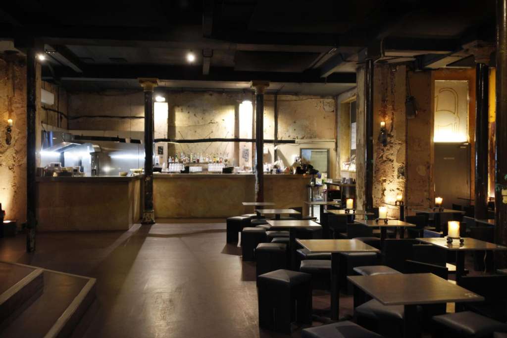 Industrial venue interior with bar counter, low seating areas, concrete floors, and warm accent lighting on exposed brick walls.