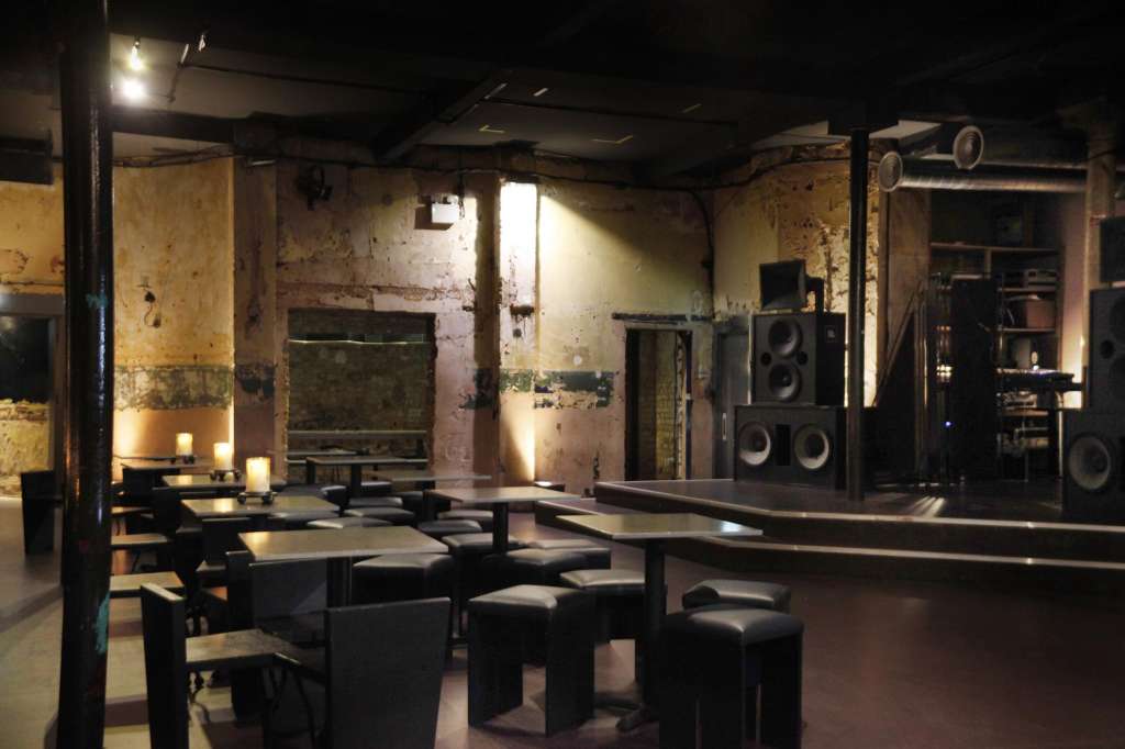 Industrial venue with exposed brick walls, black furniture, stage with speakers, and warm ambient lighting.