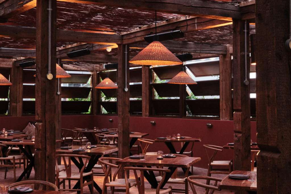 Terrace with wooden tables and chairs under woven cone pendant lights and dark timber pergola.