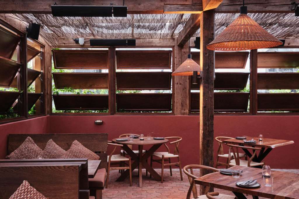 Terrace with wooden tables, woven pendant lamps, red walls, and retractable shade screens overlooking greenery.