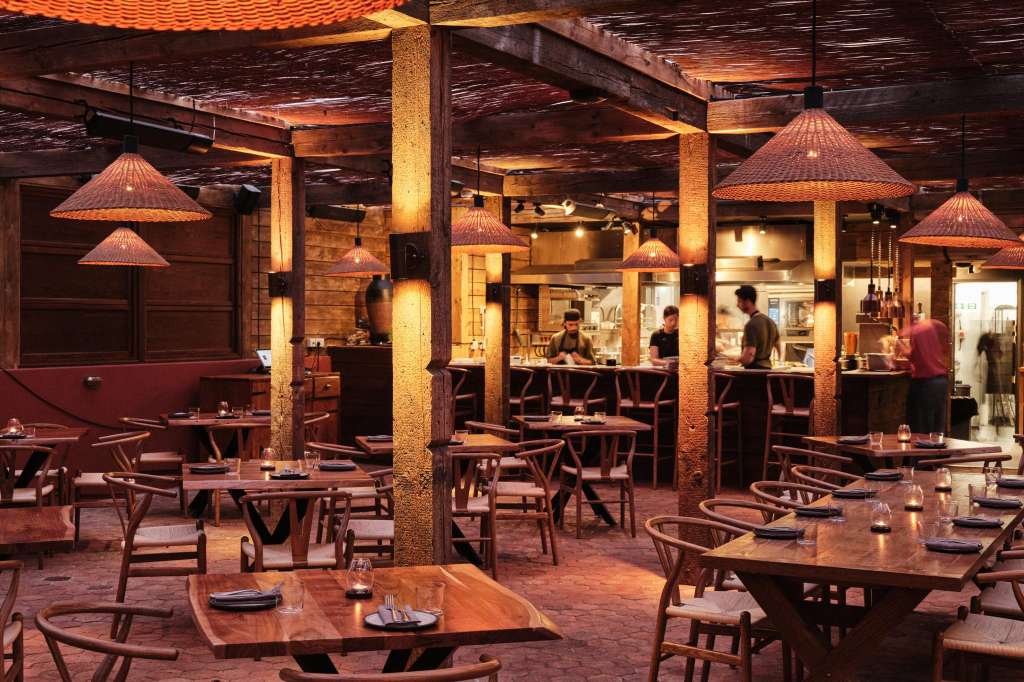 Restaurant dining area with wooden tables, mid-century chairs, copper pendant lights, and exposed brick columns.