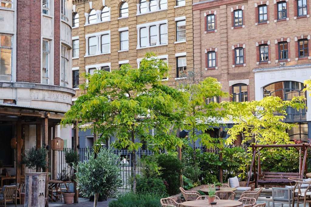 Terrace with wooden seating, potted plants, and flowering shrubs overlooking historic brick buildings.