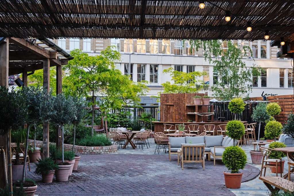 Terrace with slatted pergola, potted plants, lounge seating, and dining tables overlooking urban courtyard.