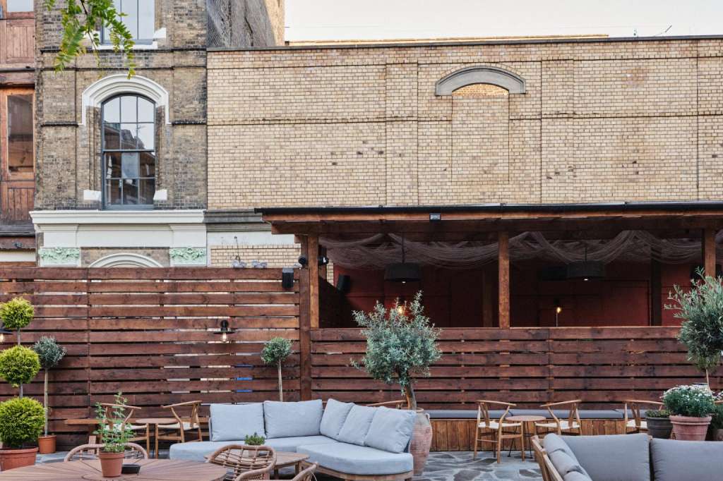Terrace with lounge seating, round tables, potted plants, and brick building backdrop under clear sky.