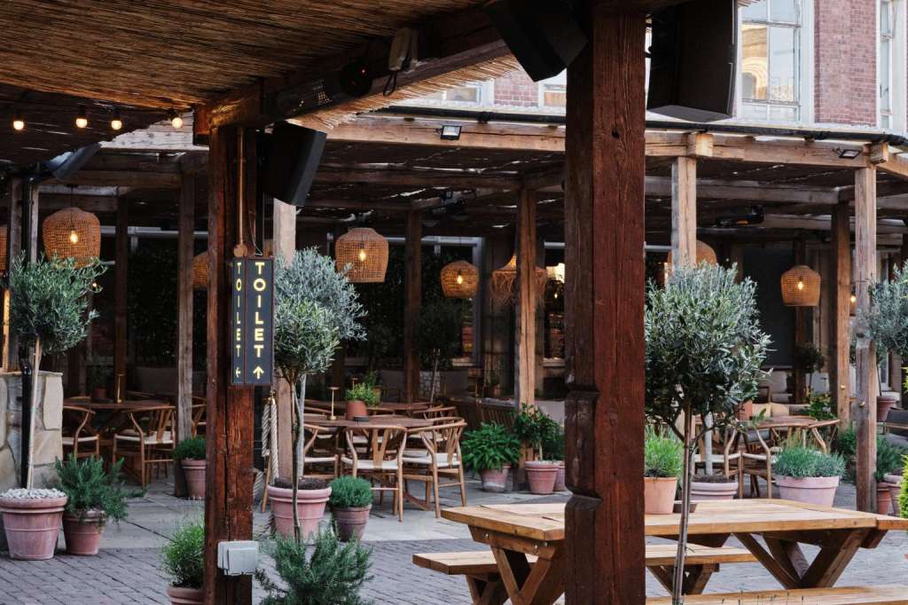 Terrace with wooden pergola, string lights, potted plants, and wooden tables set for dining.