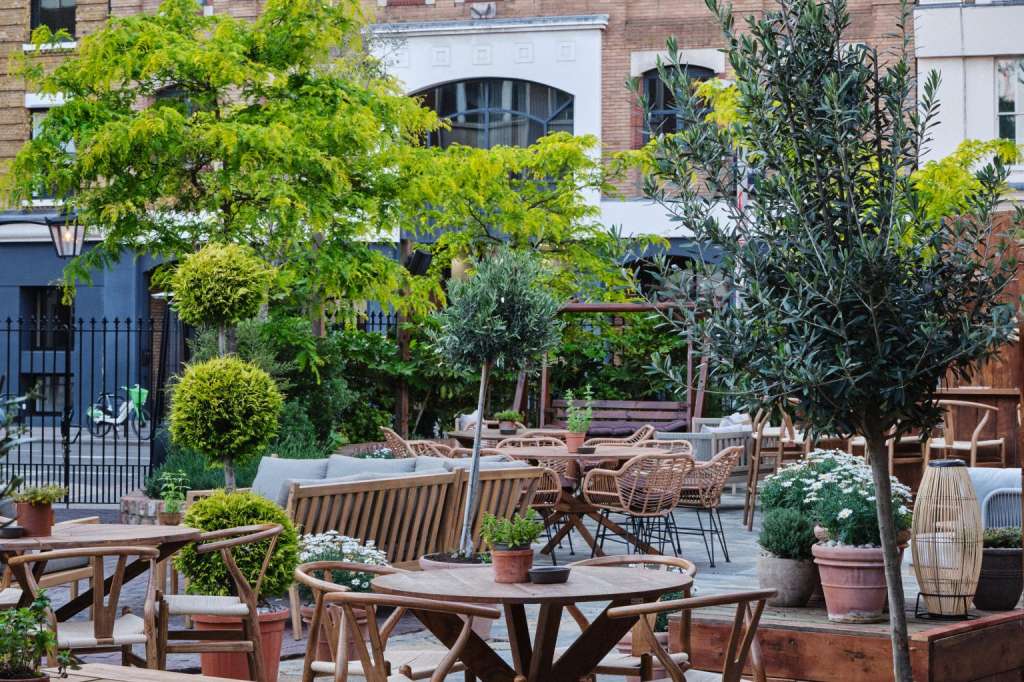 Terrace with wooden tables and chairs, lush green trees, potted plants, and brick building backdrop.