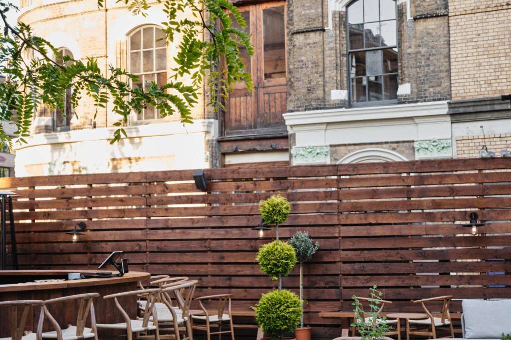 Terrace with round tables, woven chairs, potted plants, and wooden privacy fence against brick building.