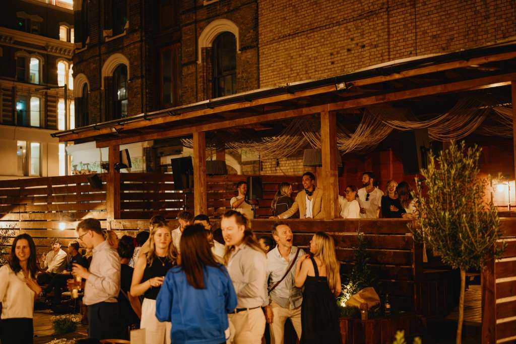 Outdoor terrace with wooden pergola, string lights, and guests mingling at an evening event.