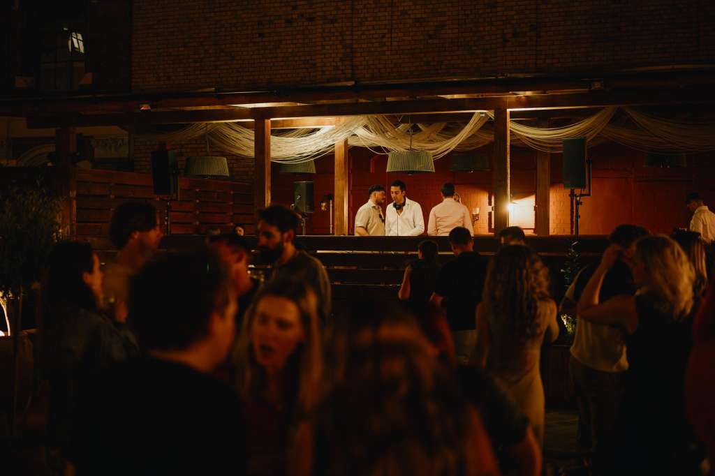Outdoor terrace at night with guests mingling, bar staff behind counter, warm lighting and draped fabric overhead.