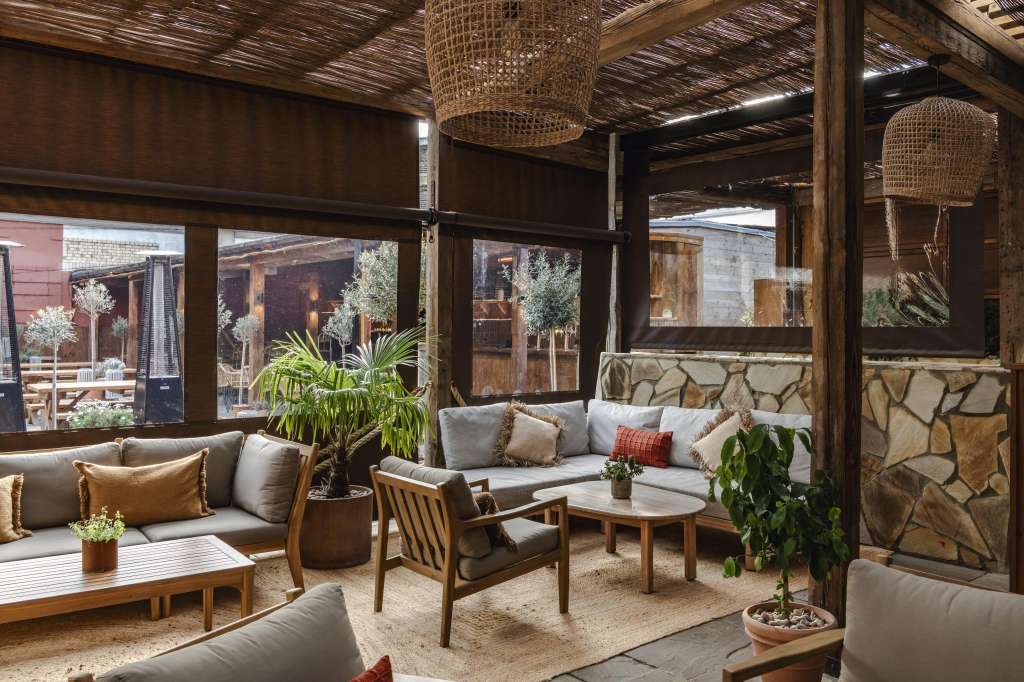 Terrace with lounge seating, woven pendant lights, stone walls, and garden views through large windows.