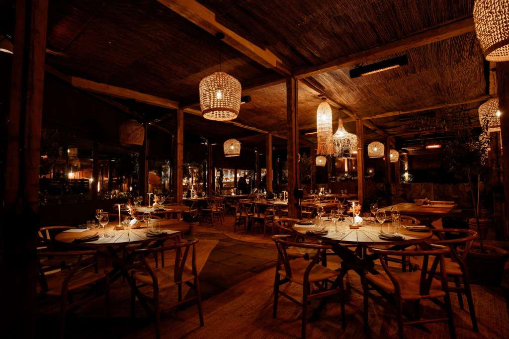 Terrace dining area set with round tables, place settings, and woven pendant lighting at night.