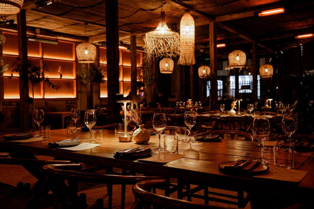 Restaurant dining room set for a formal dinner with long wooden table, place settings, wine glasses, and woven pendant lighting.