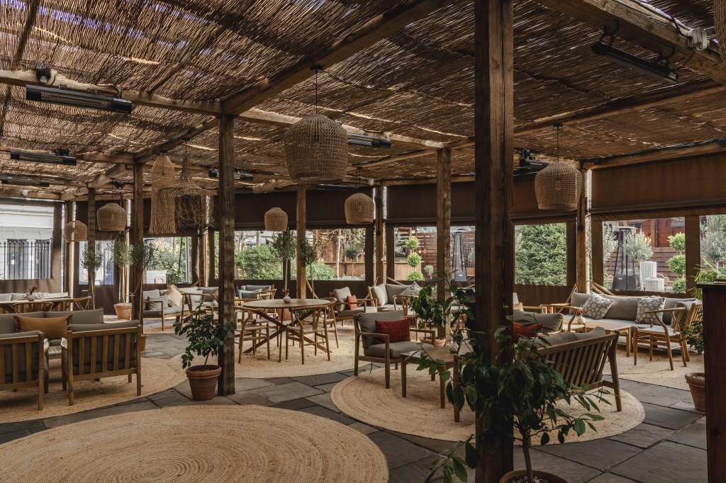 Covered terrace with wooden furniture, woven pendant lights, potted plants, and garden views through large windows.