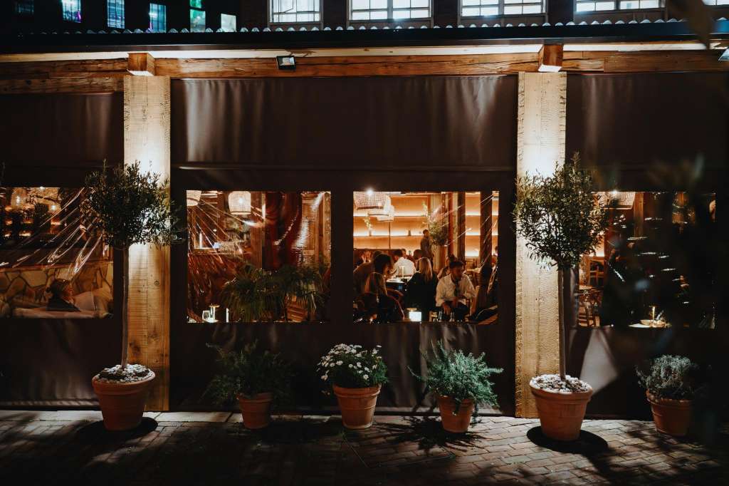 Terrace with retractable awning, potted plants, and guests visible through windows at evening.