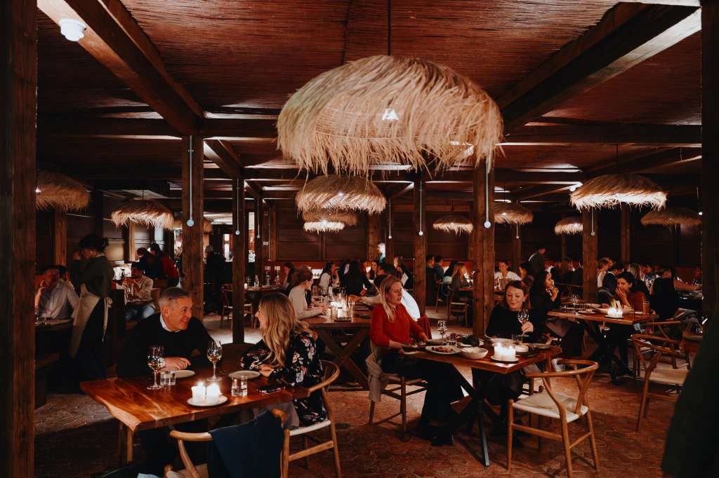 Restaurant with wooden beams and rattan pendant lights, guests seated at tables for dinner service.