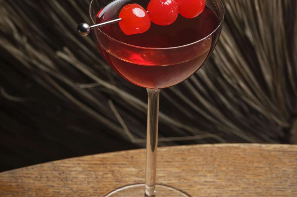 Close-up of a red cocktail with cherry garnish on a wooden surface against striped backdrop.