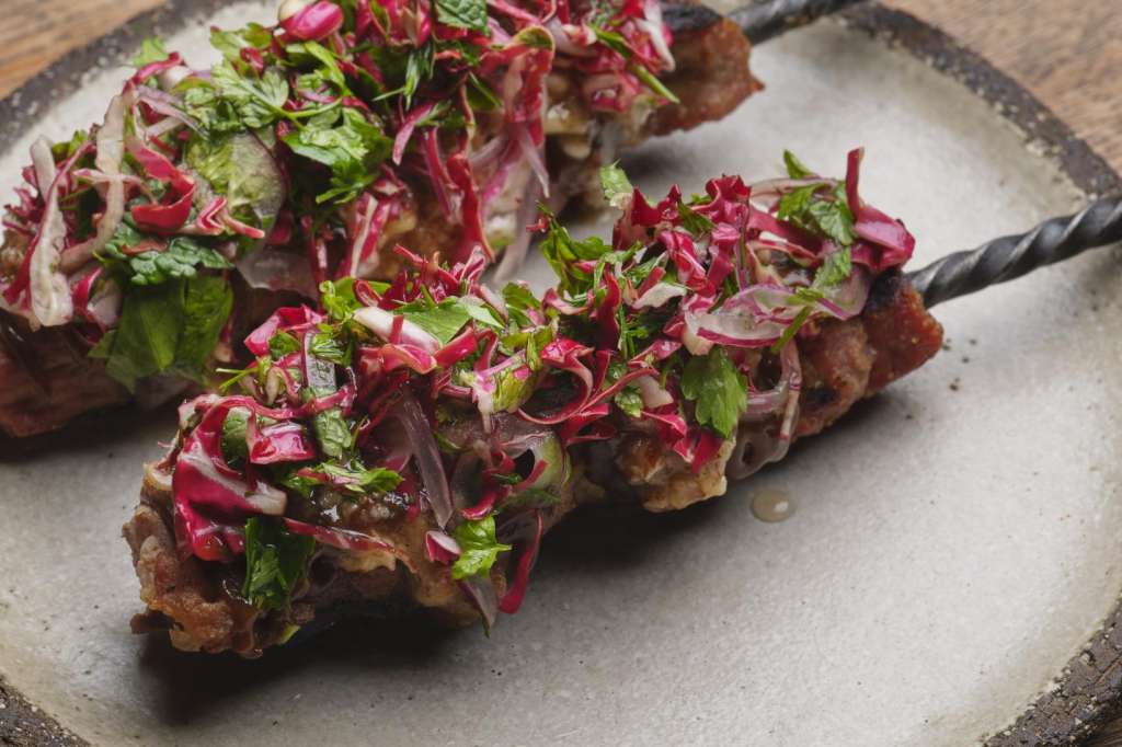 Plated meat dish with beetroot and herb garnish on ceramic plate with metal utensils.