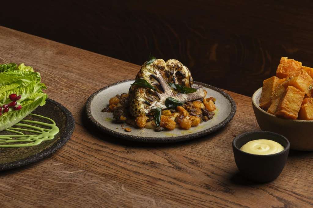 Wooden table with plated food: grilled lettuce, roasted cauliflower, fried potatoes, and sauce.