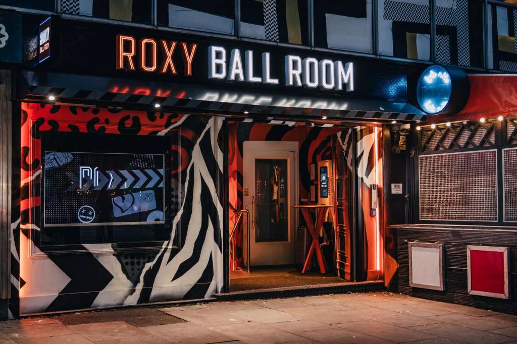 Exterior storefront of Roxy Ball Room venue at night with neon signage, red and black geometric branding, and illuminated entrance.