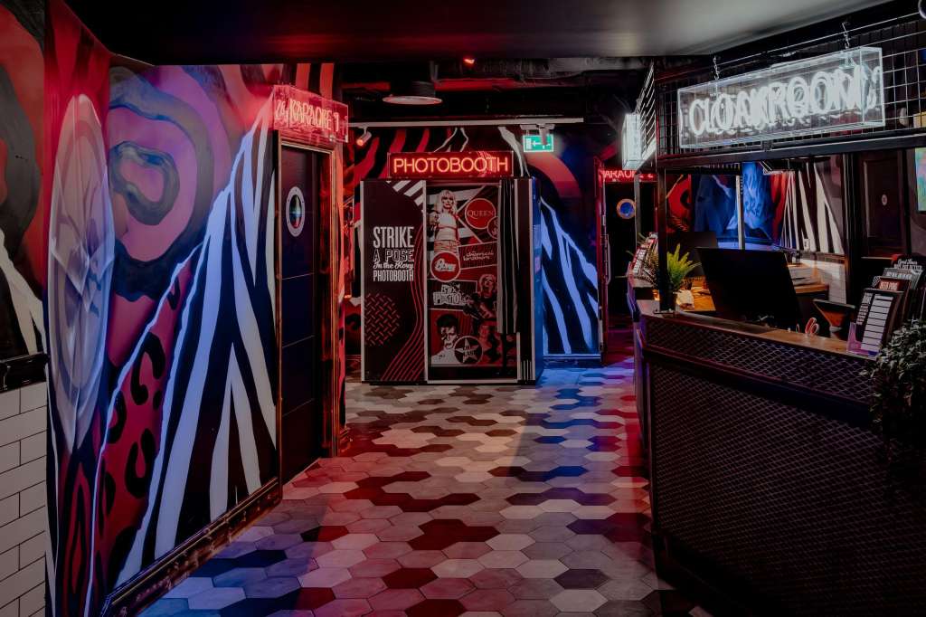 Neon-lit corridor with red and blue signage, photobooth entrance, and geometric floor tiles. Modern entertainment venue interior.