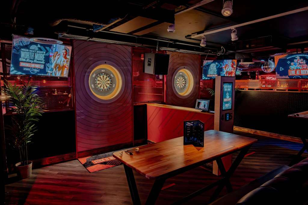 Entertainment space with dartboards, wooden table, red lighting, and branded signage on walls.