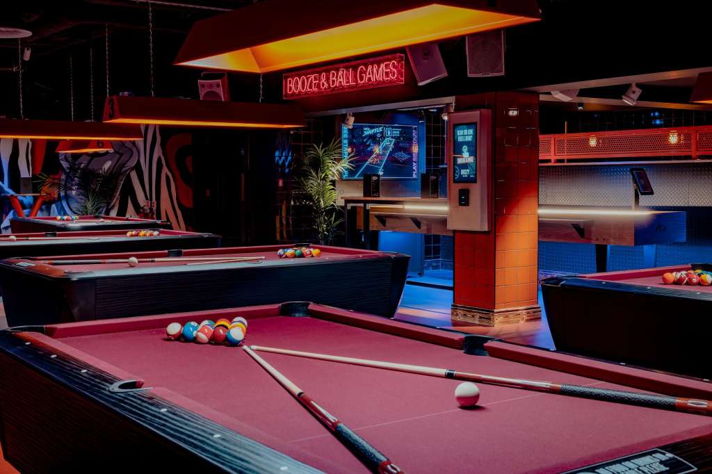 Pool tables with colorful balls and cues under neon lighting, booze and ball games signage visible overhead.