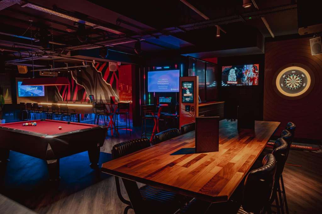 Entertainment space with pool table, wooden dining table, dartboard, and neon-lit bar area with gaming machines.