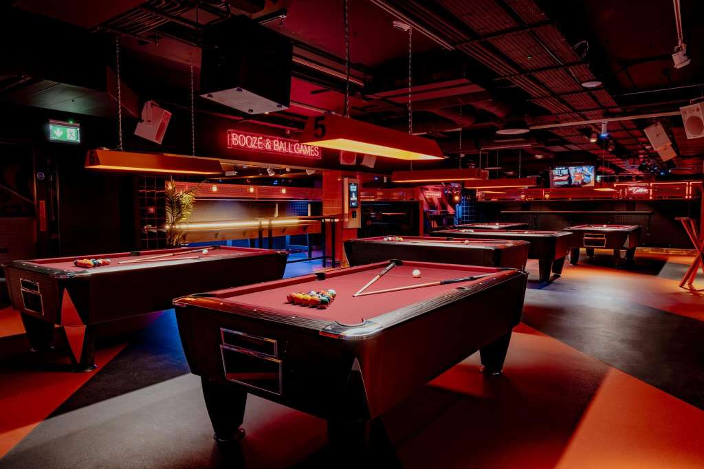 Gaming hall with multiple pool tables, red and orange lighting, neon signage, and industrial ceiling.