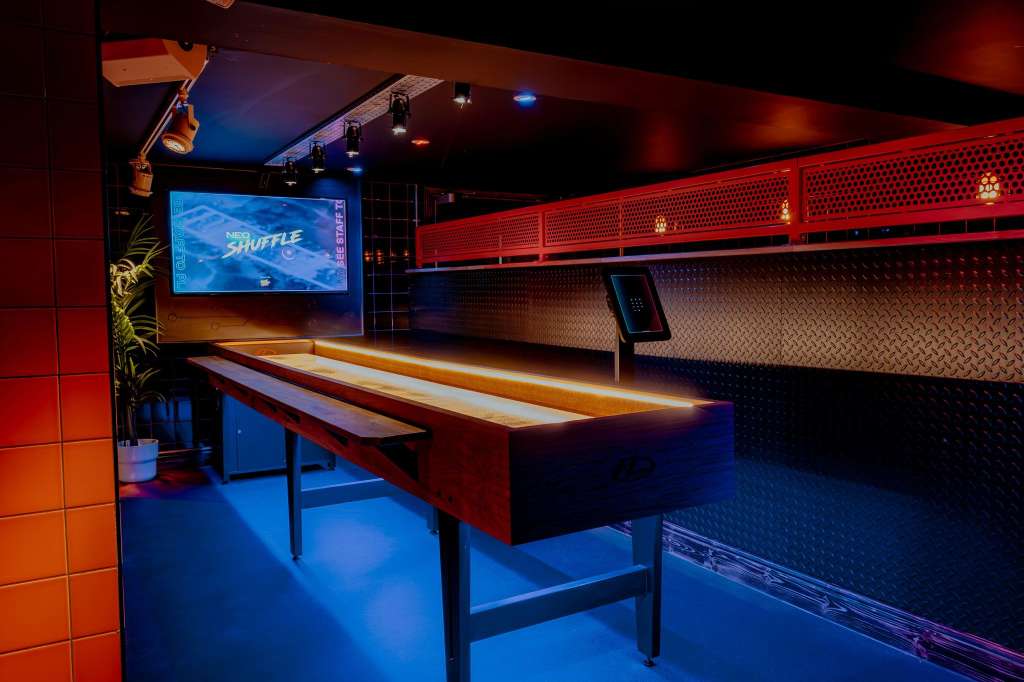 Gaming hall with illuminated shuffleboard table, blue and red ambient lighting, and video screen on wall.