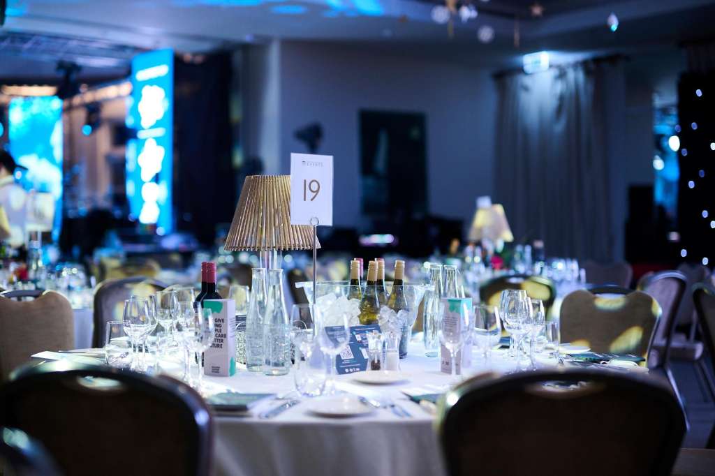 Formal dining table set with white linens, glassware, place cards, and gold table lamps under blue ambient lighting.