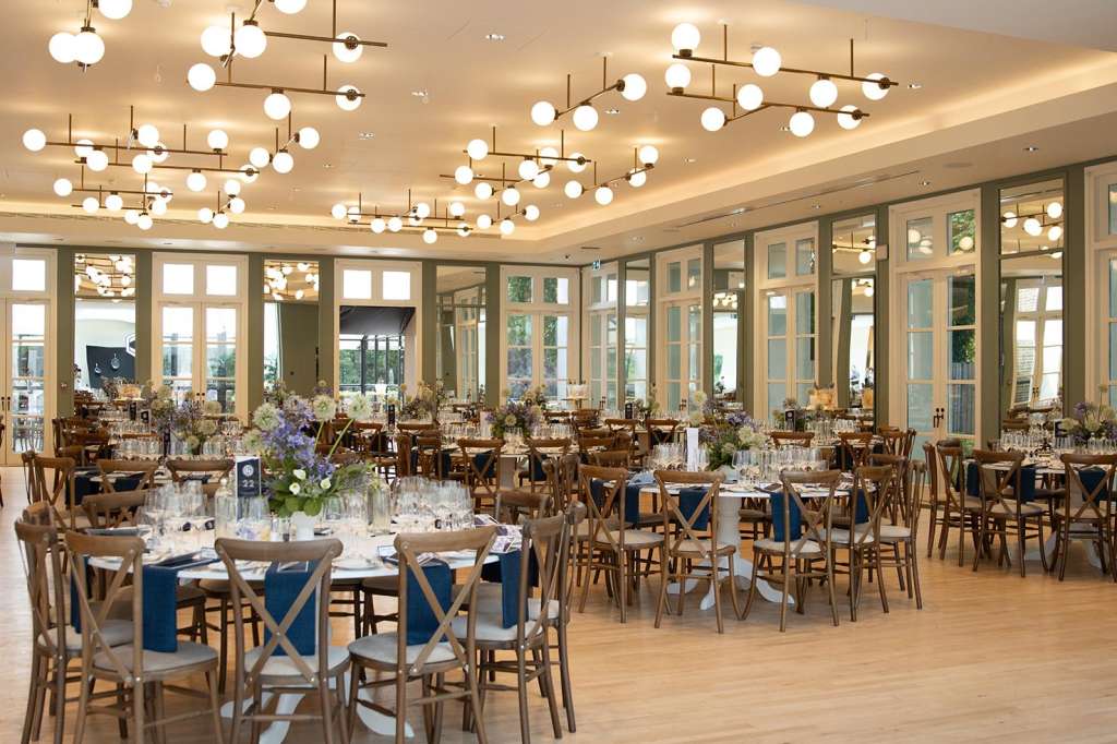 Palm Court set up for a seated dinner with round tables, wooden chairs, blue napkins, and modern globe pendant lighting.