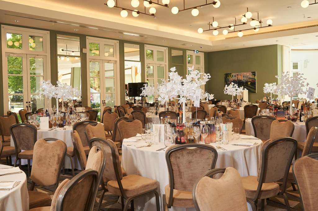 Elegant dining room with round tables, white linens, upholstered chairs, and white floral centerpieces under modern pendant lighting.
