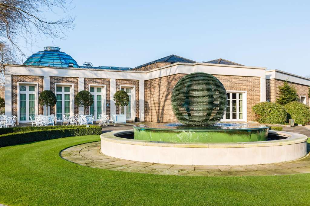 Exterior courtyard with manicured lawn, circular water feature, and brick pavilion with white chairs visible.