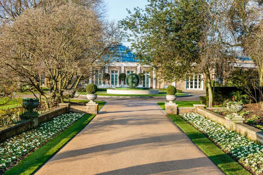 Exterior pathway lined with flowering shrubs leading to a classical building with blue dome and manicured gardens.