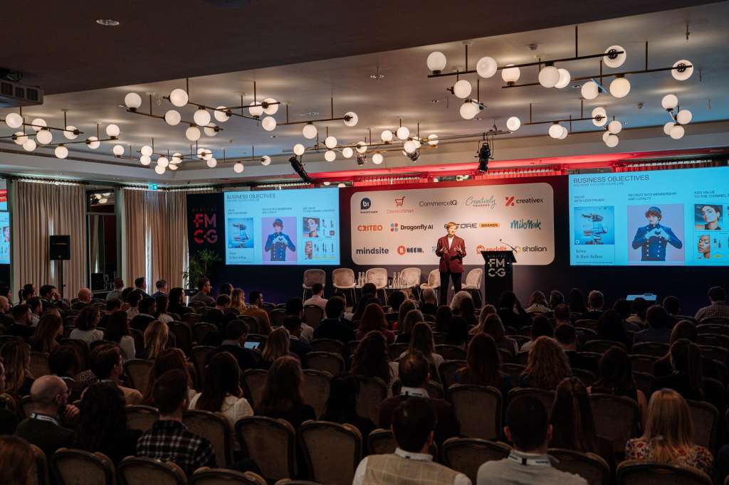 Large conference room with seated audience facing stage, multiple screens displaying business content, and modern pendant lighting.