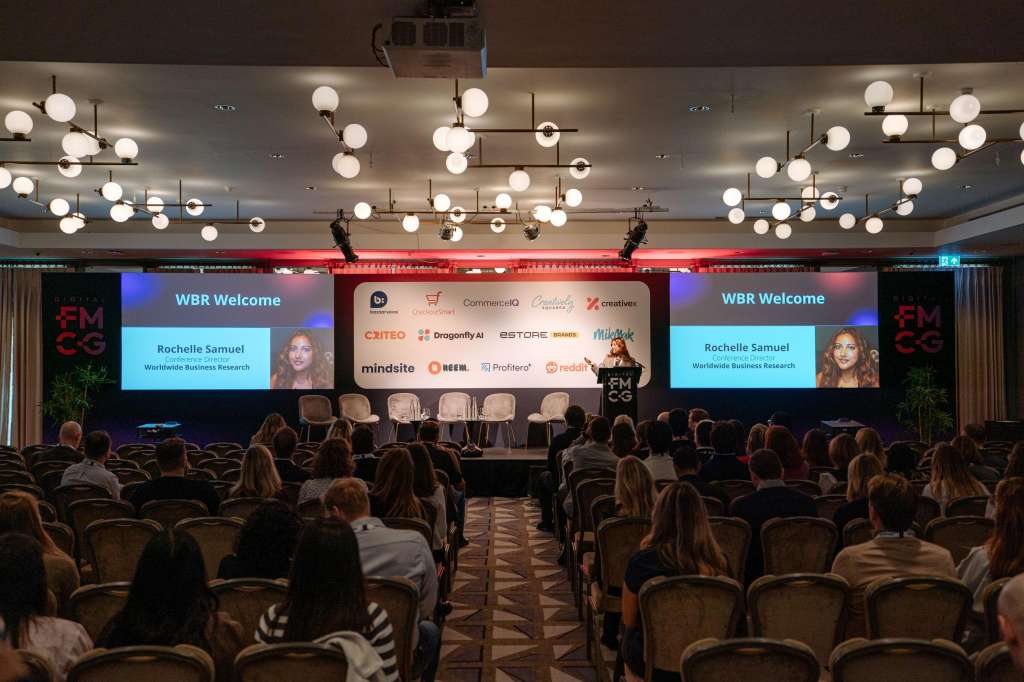 Conference room with rows of seated attendees facing a stage with multiple projection screens and sponsor logos.