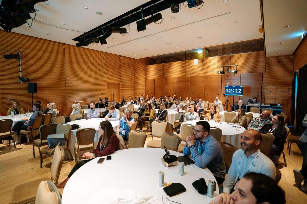 Modern wood-paneled conference room with round tables, seated attendees, professional lighting rig, and digital display.