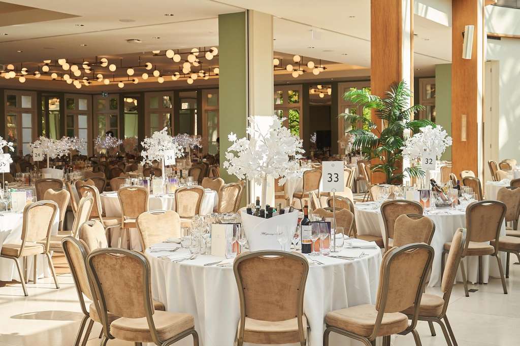 Ballroom set for seated dining with round tables, white linens, upholstered chairs, white floral centerpieces, and pendant lighting.
