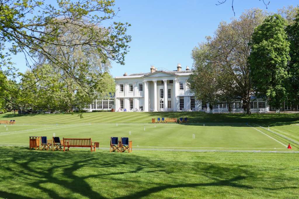 Manicured lawn with croquet setup, wooden seating, and classical white building with columns in background.
