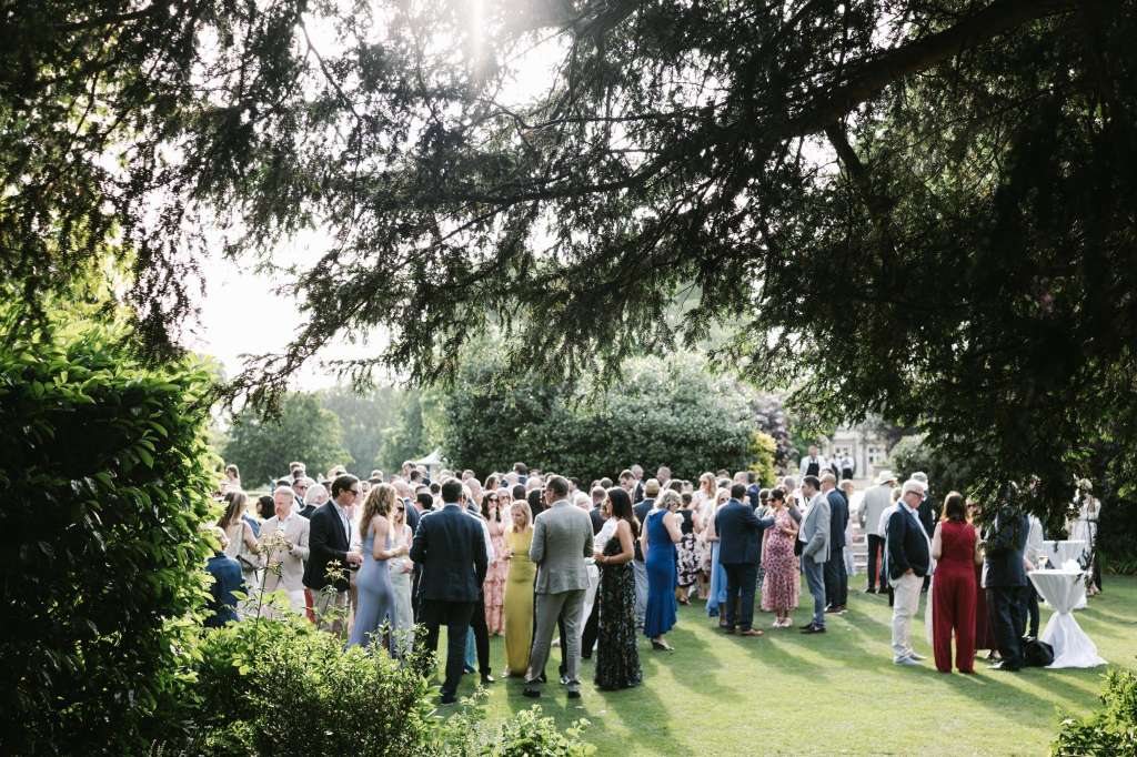 Outdoor garden reception with guests mingling under mature trees on manicured lawn.