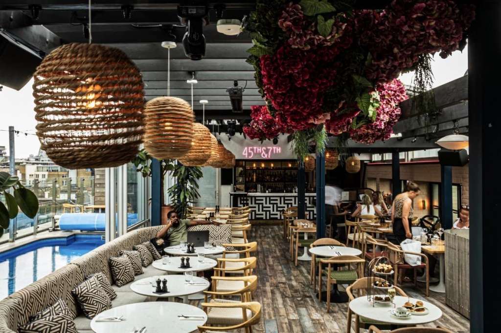 Rooftop dining space with round tables, woven pendant lights, burgundy floral installations, and city views through floor-to-ceiling windows.