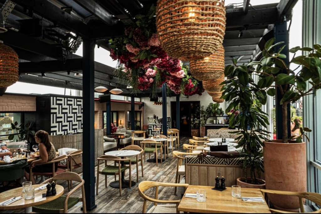 Rooftop restaurant with wooden tables, woven pendant lights, burgundy floral installations, and potted plants under black pergola.