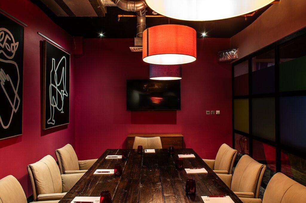 Meeting room with burgundy walls, long dark wood table, beige chairs, red pendant lighting, and neon wall art.