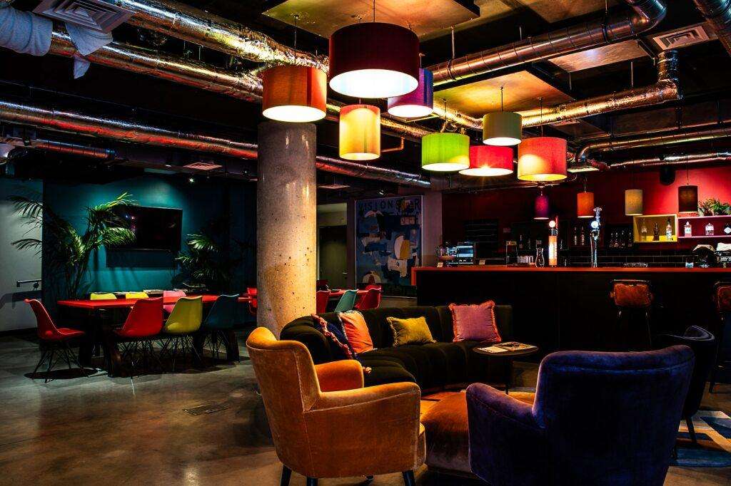 Modern event space with colorful pendant lighting, bar counter, lounge seating, and industrial exposed pipes.