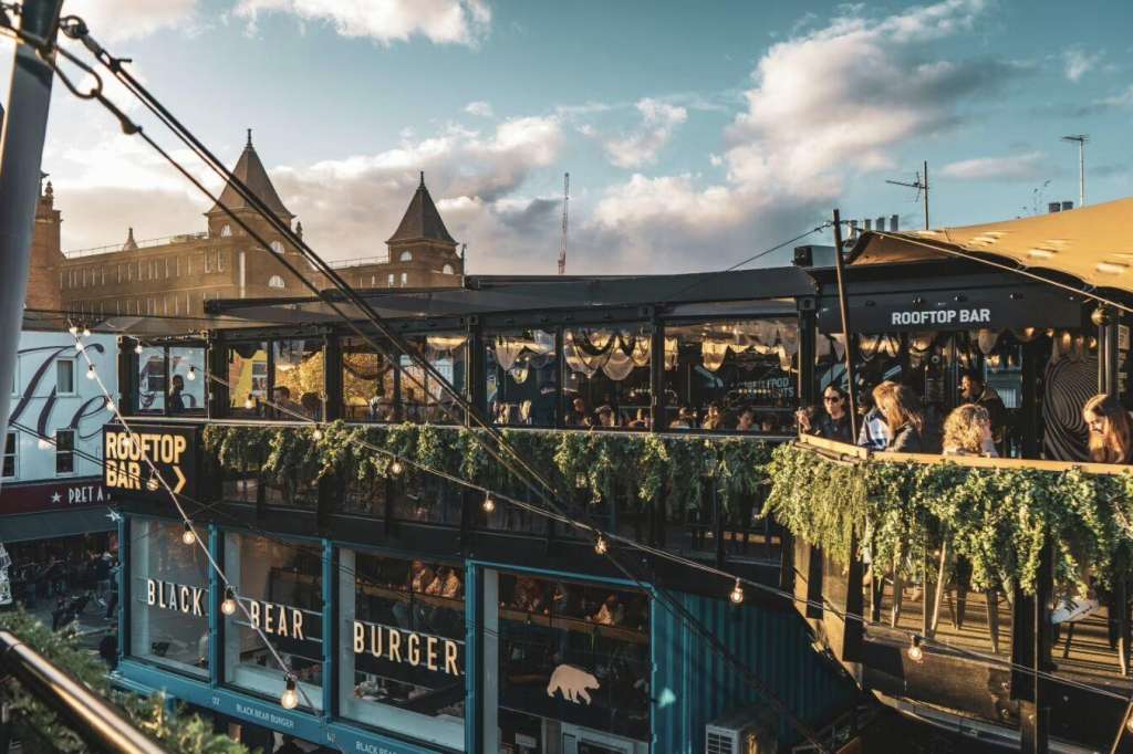 Rooftop bar with crowds, hanging greenery, string lights, and urban skyline backdrop in golden hour sunlight.
