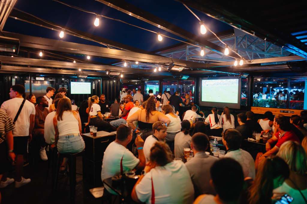 North Terrace at night with string lighting, multiple screens, and a crowd of guests mingling and standing.