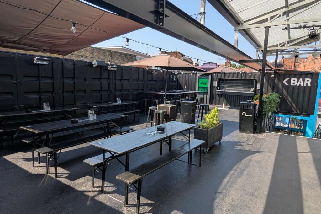 Outdoor terrace with picnic tables, retractable shade canopy, and bar signage under clear blue sky.