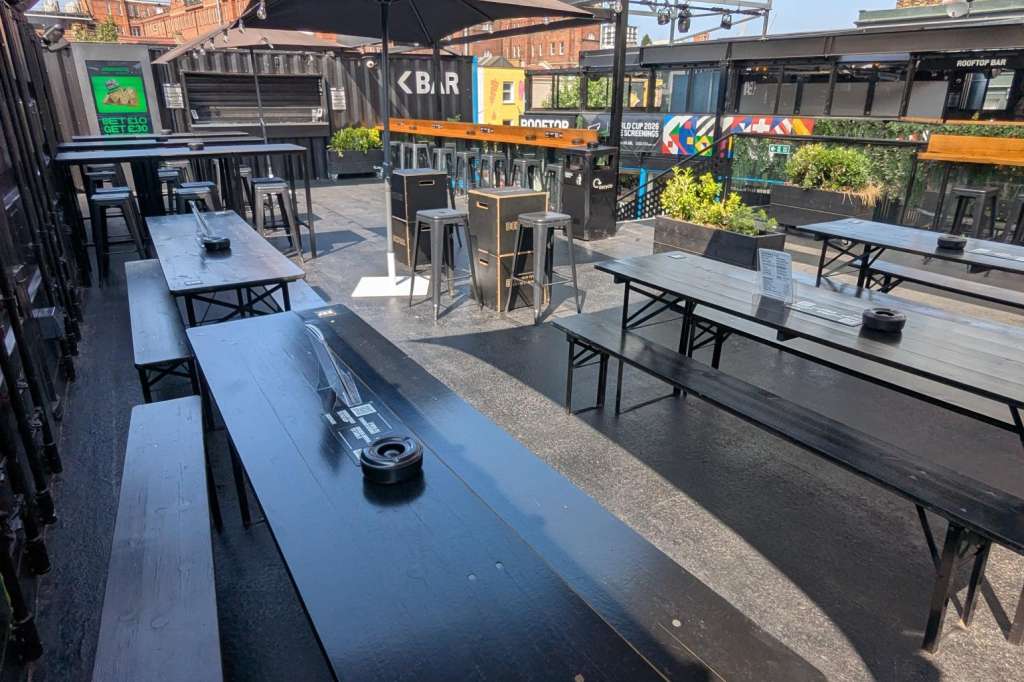 Outdoor terrace with long tables, benches, black umbrellas, and bar counter under steel canopy.