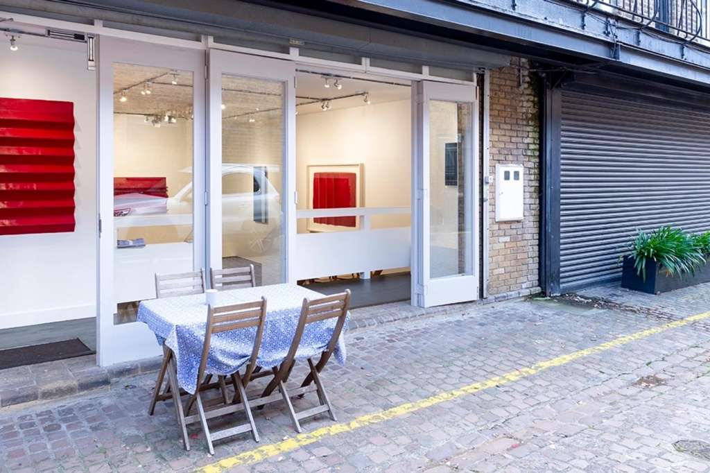 Contemporary gallery storefront with red abstract artworks displayed behind glass, outdoor seating area with table and chairs.