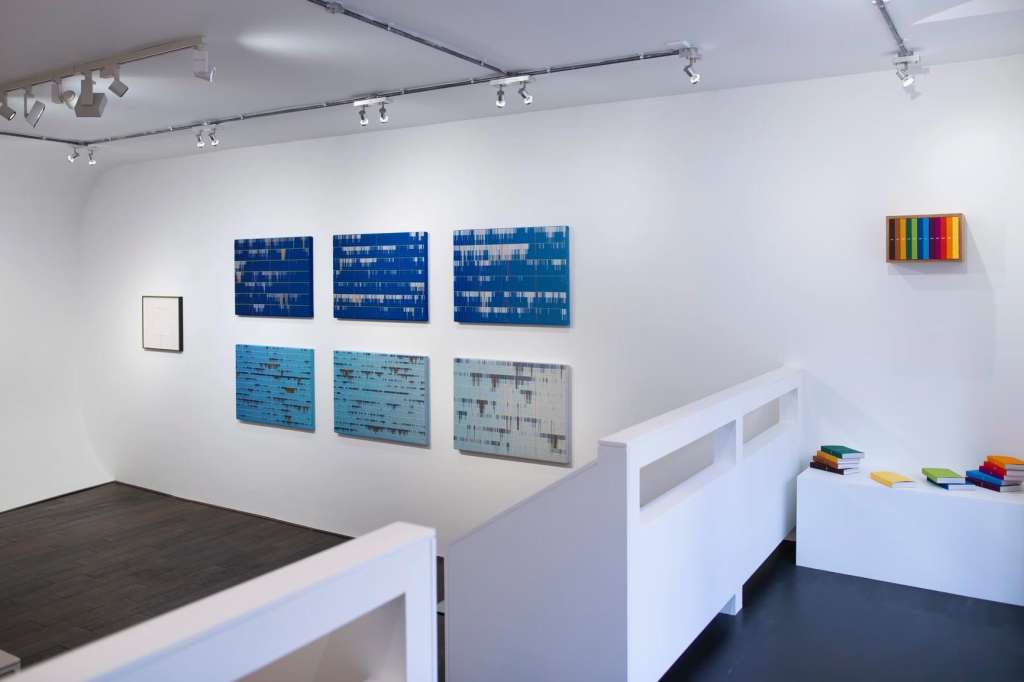 Gallery space with blue abstract artworks on white walls, track lighting, and white display pedestals.